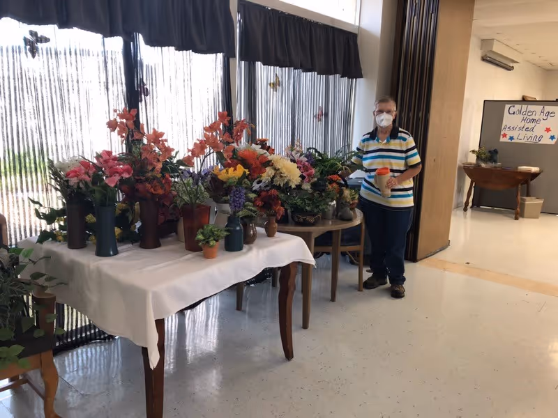 An elderly person wearing a striped shirt and face mask stands next to tables covered with various colorful potted flowers and plants inside a well-lit room. A sign on the wall in the background reads 'Golden Age Home Assisted Living.'