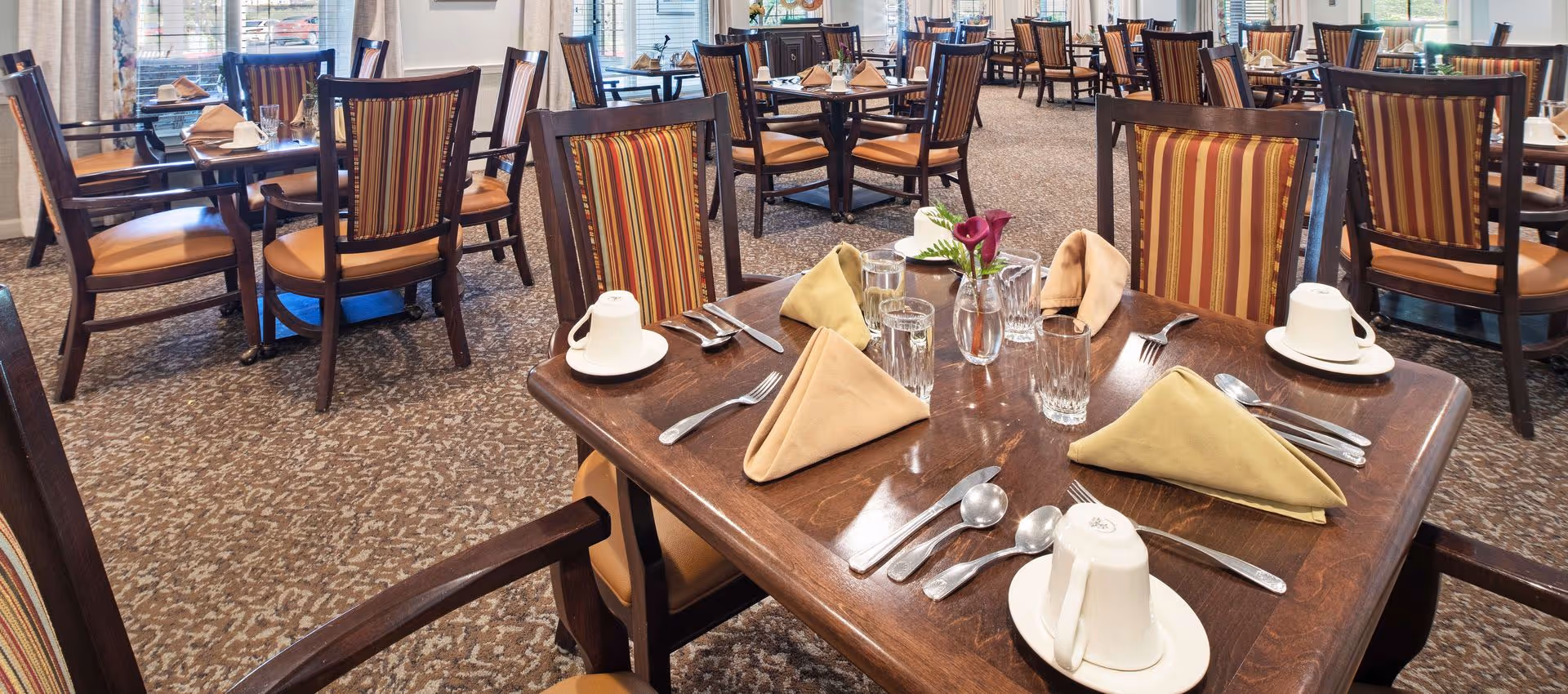 A dining room with multiple wooden tables and chairs featuring striped upholstery. Each table is set with folded beige napkins, glasses of water, white coffee cups on saucers, silverware, and a small vase with a purple flower. Large windows with blinds and curtains allow natural light into the room.
