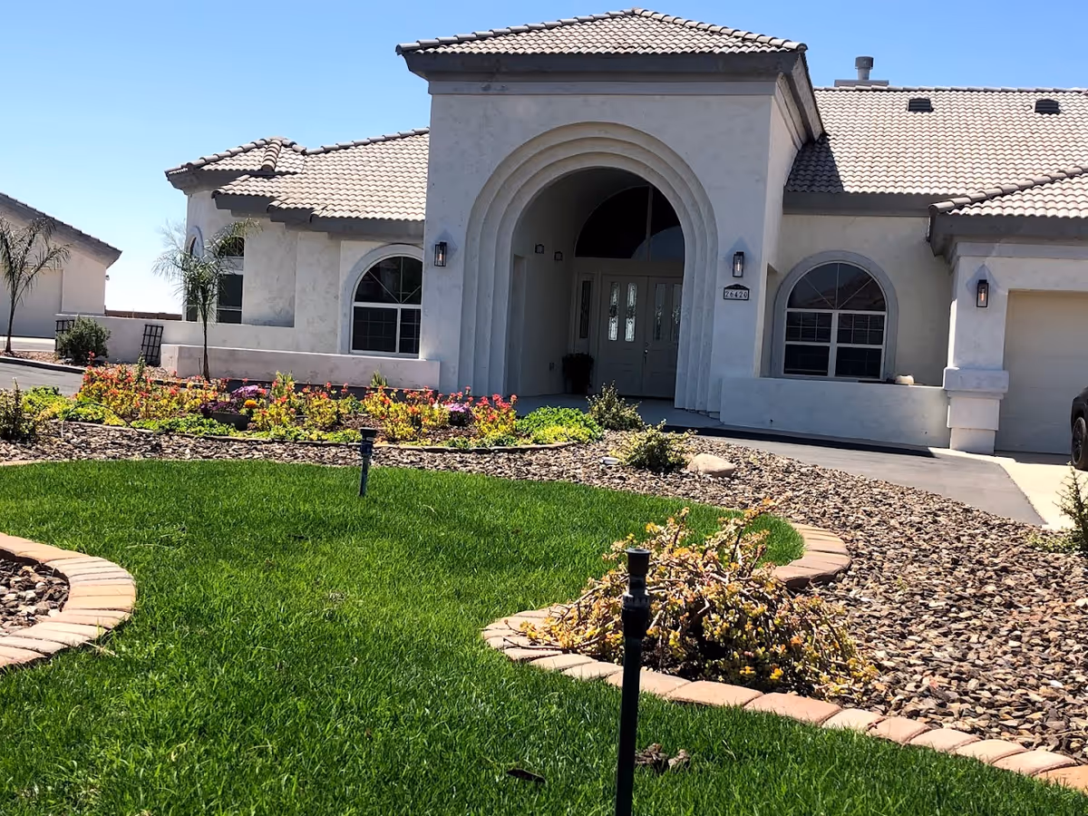 Front exterior view of a single-story building with a tiled roof, arched entrance, and large windows. The building is surrounded by a well-maintained garden with green grass, flower beds, and small palm trees under a clear blue sky.