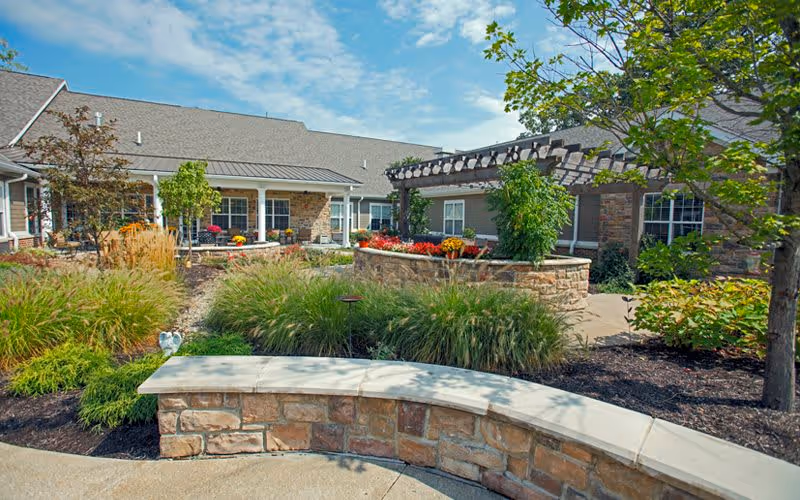 Outdoor garden area at Bickford of Carmel featuring a curved stone bench, various plants and shrubs, a pergola with climbing greenery, and a building with a covered porch in the background under a partly cloudy sky.