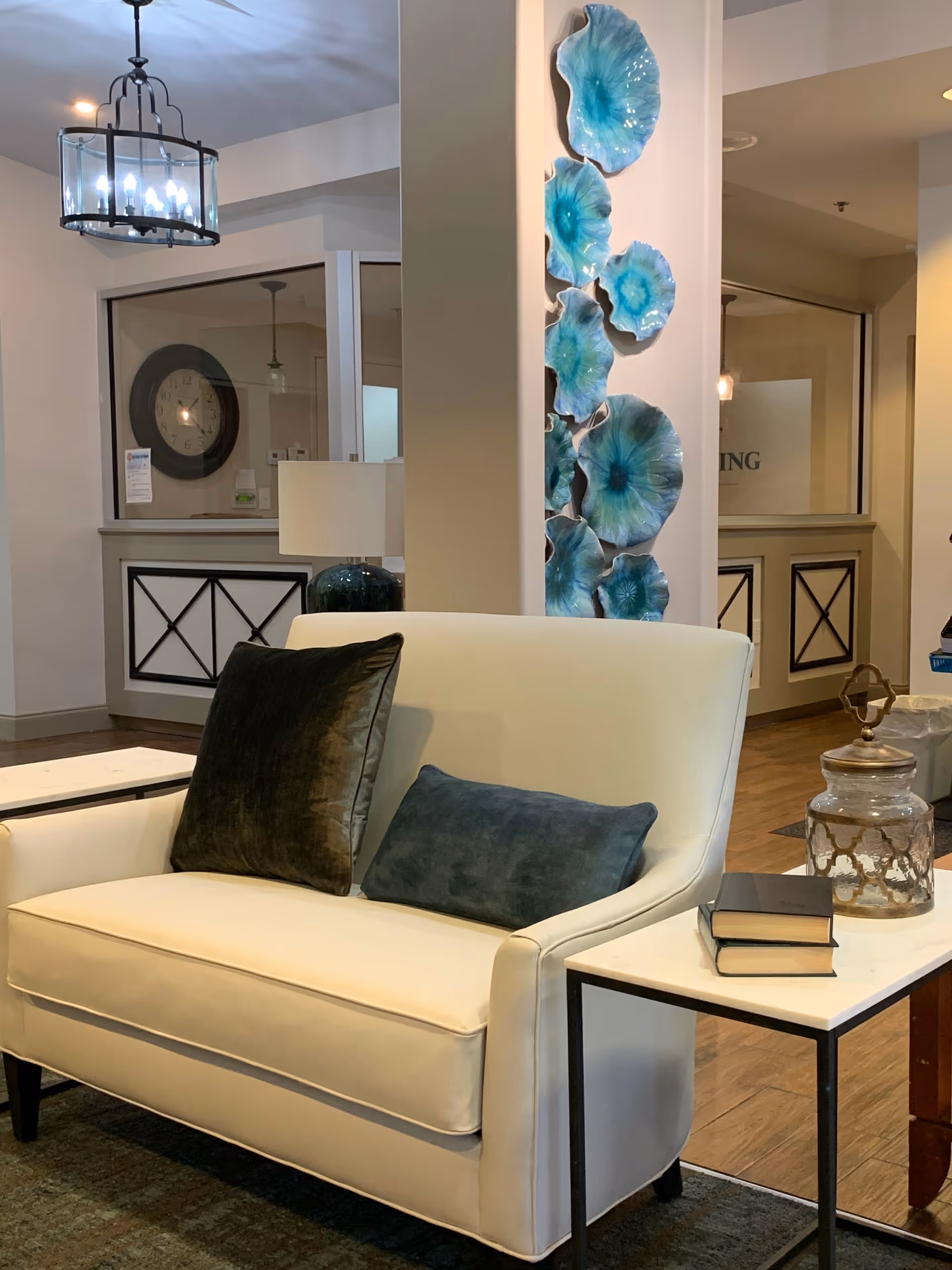 A cozy seating area in a living room with a white loveseat adorned with two dark-colored pillows. Next to the loveseat is a small white side table holding two stacked books and a decorative glass jar. Behind the loveseat is a white pillar decorated with blue and turquoise ceramic wall art resembling flowers or sea creatures. The background shows a reception or office area with a large clock on the wall and a hanging light fixture above.