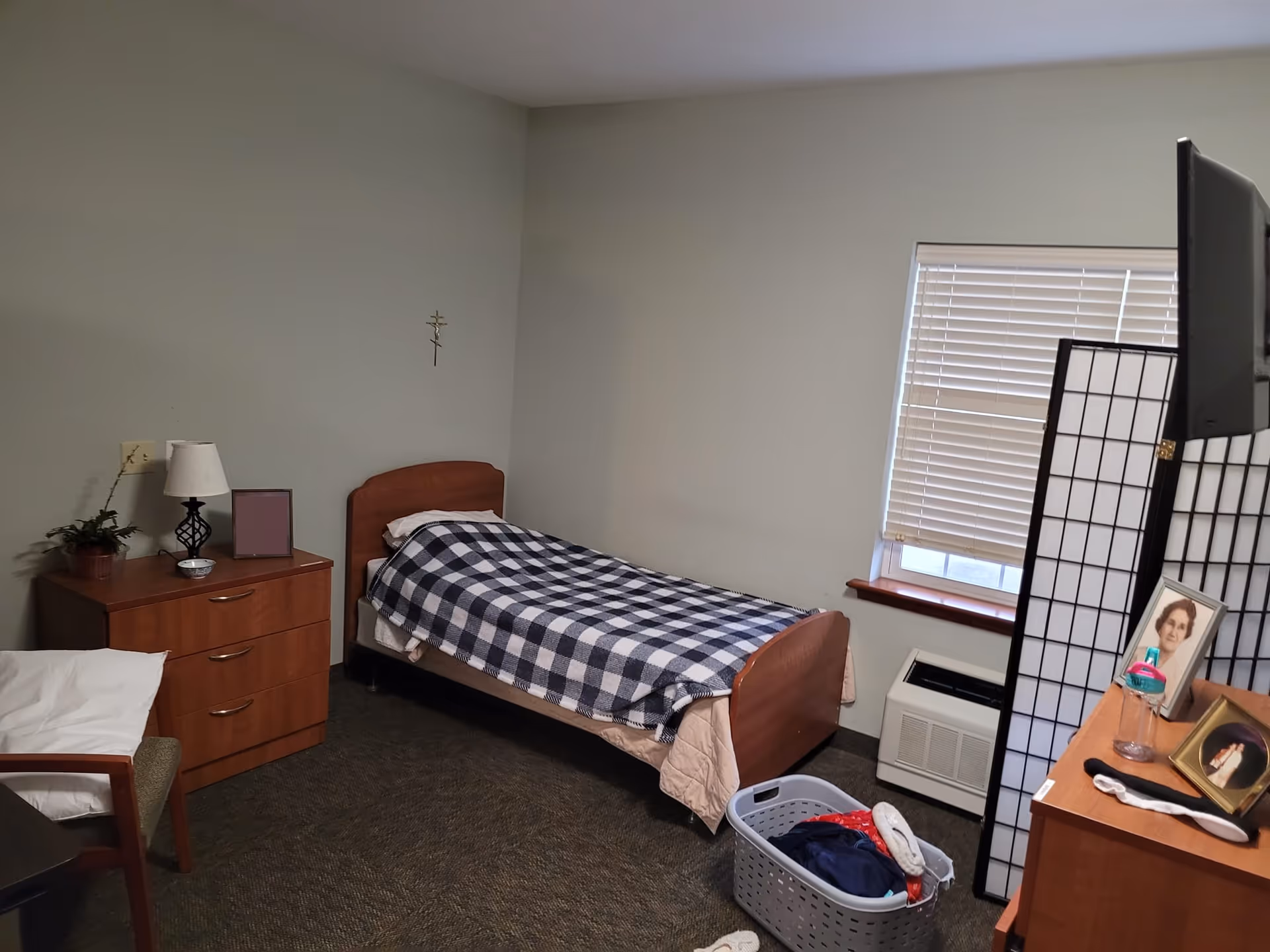 A senior living facility bedroom featuring a bed, dresser, and window blinds.