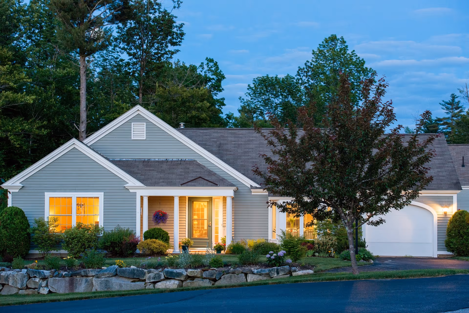 Single-story gray house front with lit windows, a porch, attached garage, and landscaped yard at dusk.
