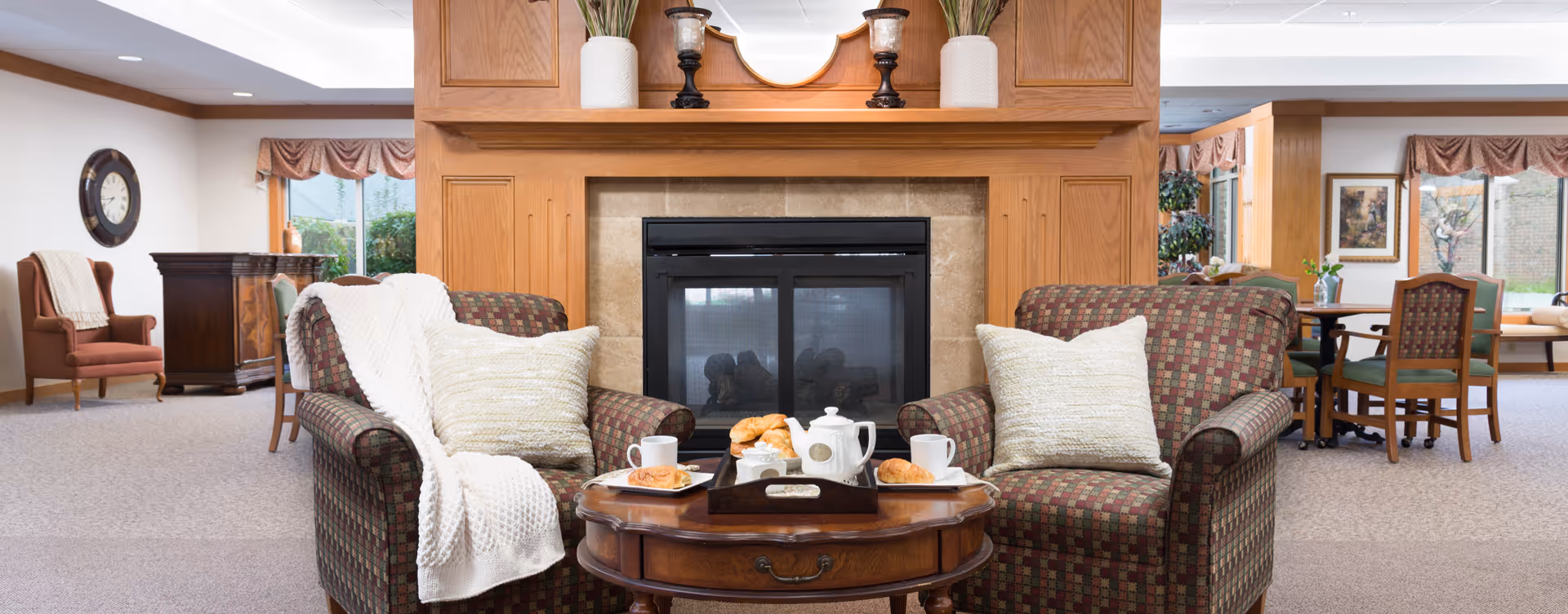 Cozy sitting area in a senior living facility with two patterned armchairs, each with a cream-colored pillow and a white knitted throw blanket. A wooden coffee table between the chairs holds a tray with a white teapot, two cups, and croissants. Behind the chairs is a fireplace with a wooden mantel decorated with vases and candle holders. The room has large windows with valances, a clock on the wall, and additional seating and tables in the background.
