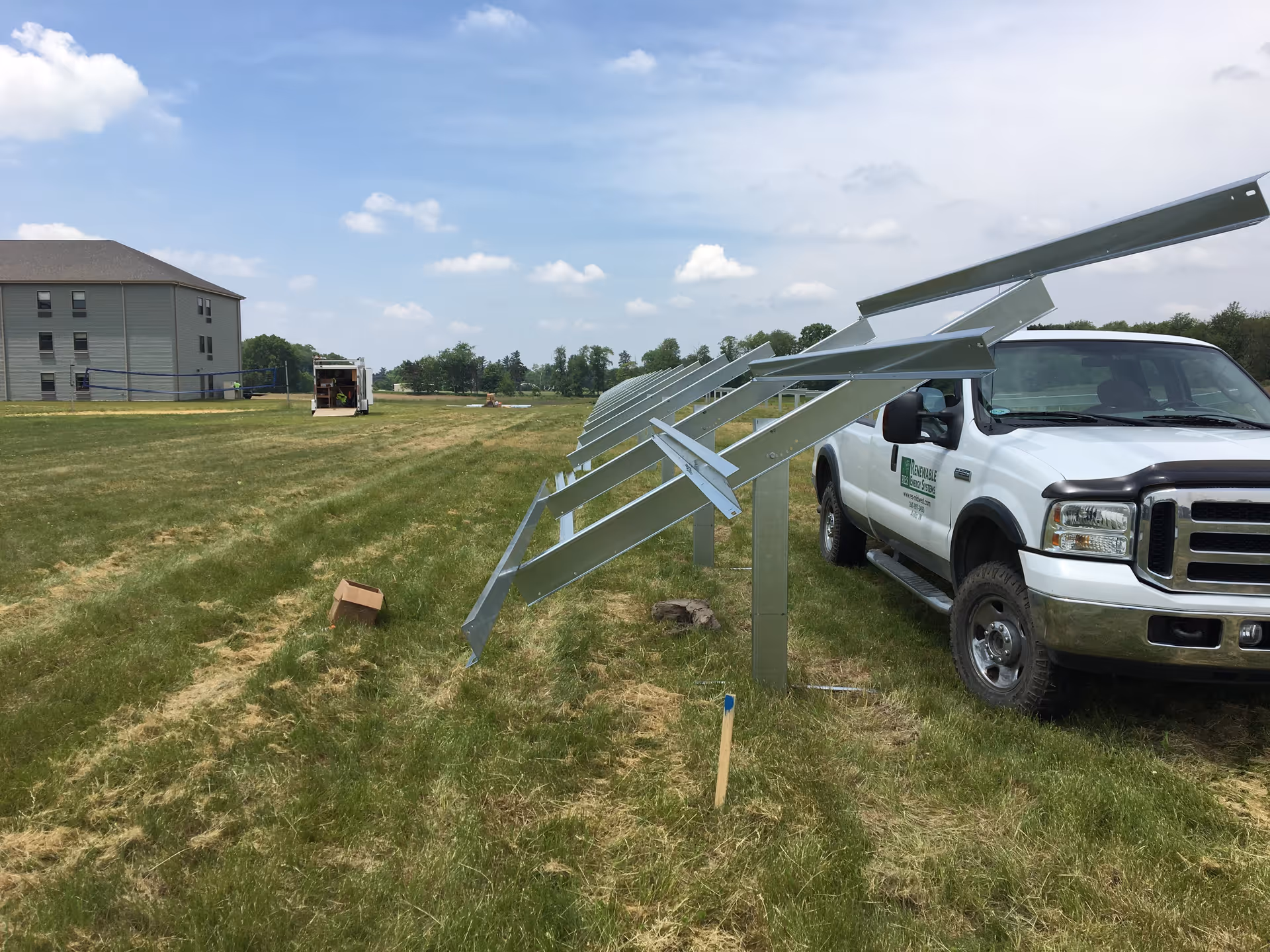 A grassy field with metal frames installed in a row, likely for solar panels, next to a white pickup truck. In the background, there is a multi-story building and a trailer. The sky is partly cloudy.