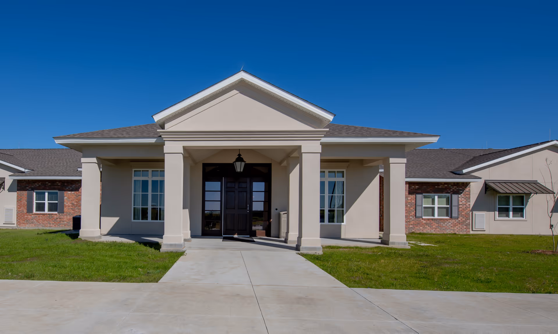 Front exterior view of a single-story assisted living facility building with a covered entrance supported by columns, large windows, and a clear blue sky.