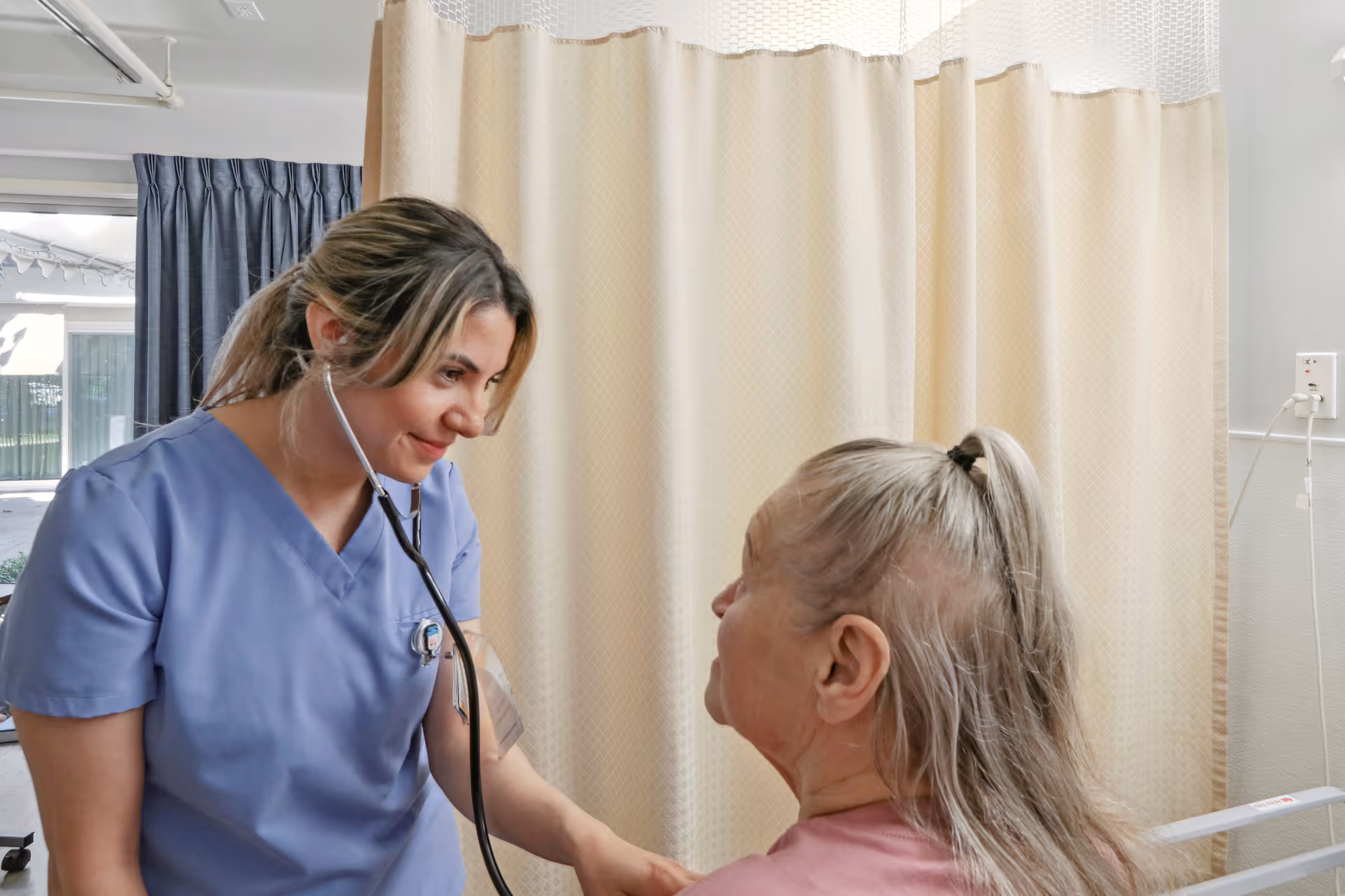 A healthcare worker in blue scrubs uses a stethoscope to check the heartbeat of an elderly woman with gray hair tied in a ponytail, seated in a medical room with beige privacy curtains in the background.