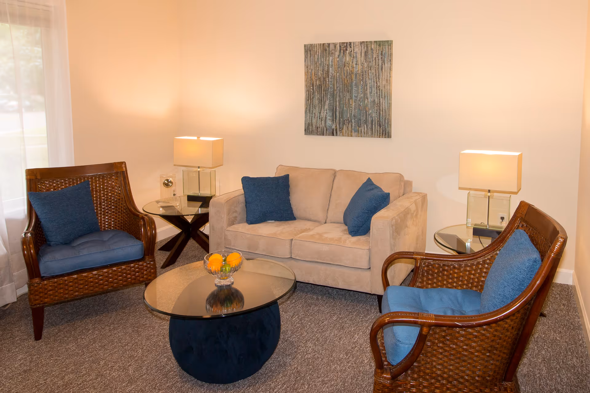A cozy living room with a beige loveseat adorned with two blue pillows, flanked by two wicker armchairs with blue cushions. There are two glass-top side tables each with a modern lamp, and a round glass coffee table with a bowl of oranges in the center. A textured abstract painting hangs on the wall above the loveseat.