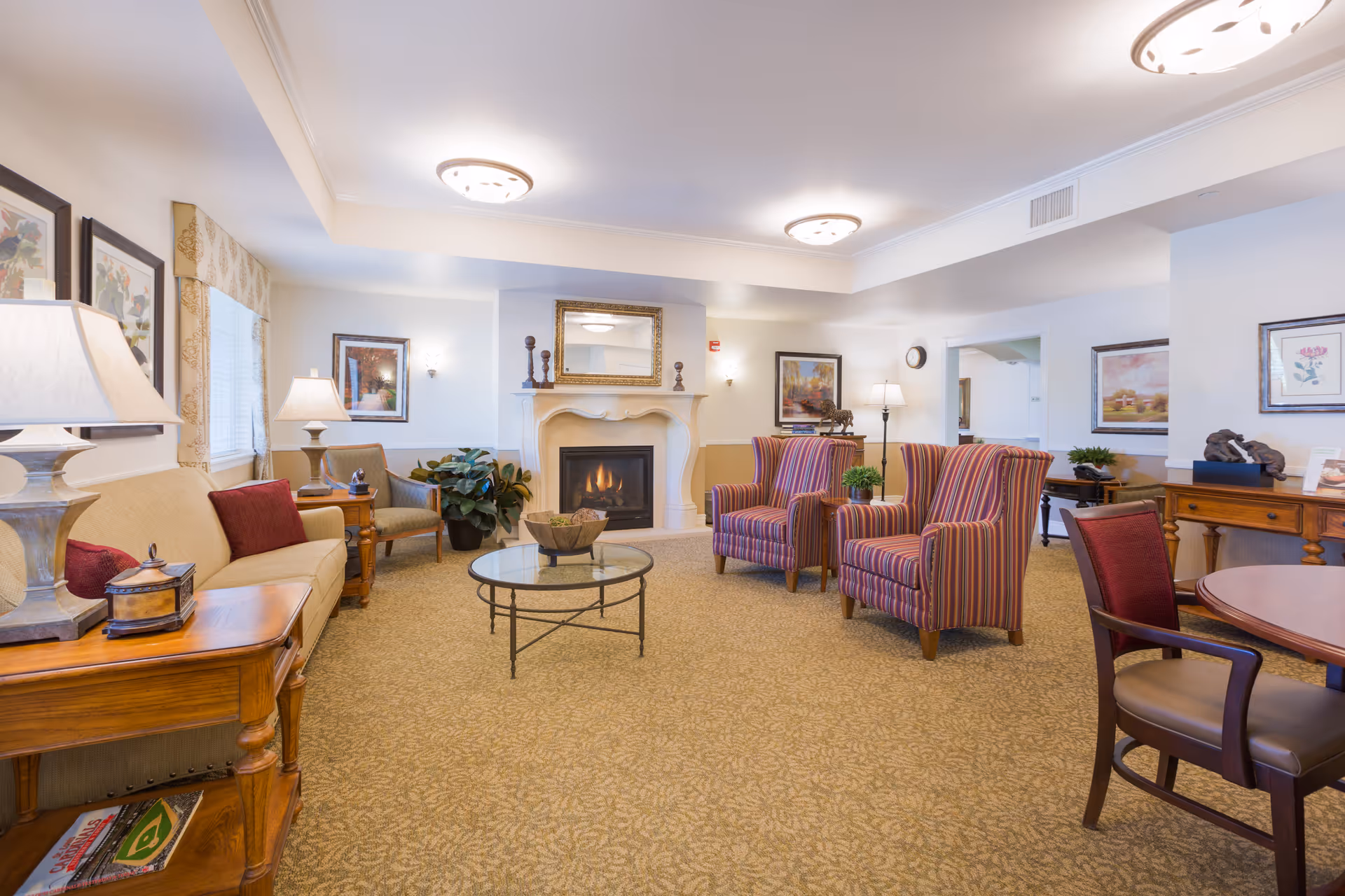 A cozy senior living facility common area with a beige sofa, two striped armchairs, a glass coffee table, and a fireplace with a mirror above it. The room is warmly lit with ceiling lights and table lamps, decorated with framed artwork and plants.