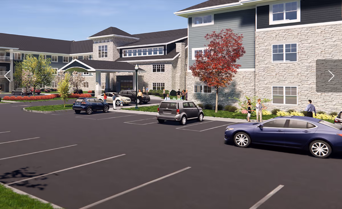 Exterior view of a senior living facility named Clarendale Of St. Peters showing a large parking lot with several cars parked and people standing near the entrance. The building has a stone facade with multiple windows and a covered entrance area. Trees and landscaped greenery surround the building.