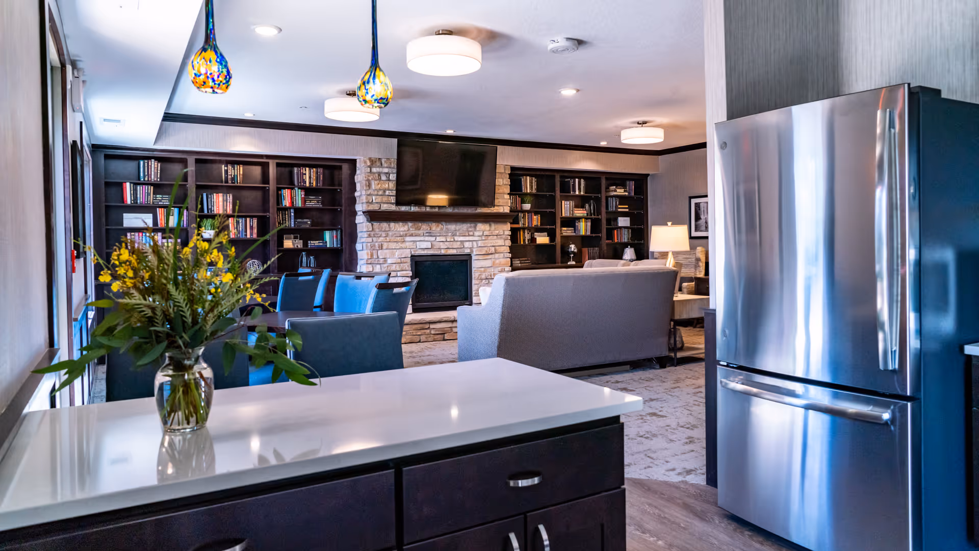 A senior living common area showing a kitchen island with a vase, stainless steel refrigerator, seating area facing a stone fireplace and built-in bookshelves.