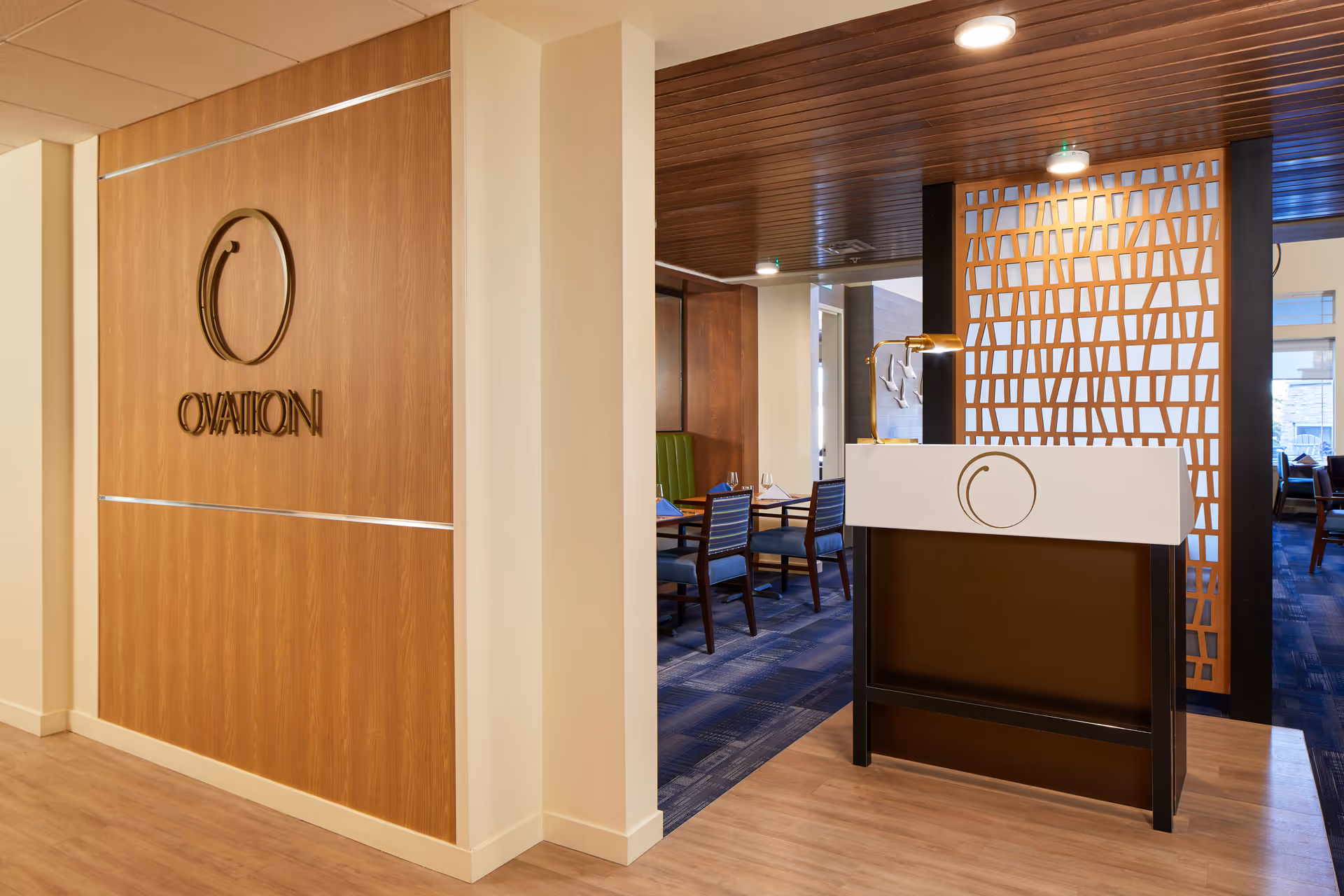 Interior view of a dining area entrance at a senior living facility named Ovation. The image shows a wooden wall with the Ovation logo and name, a reception podium with a lamp, and a glimpse into a dining room with tables, chairs, and place settings.