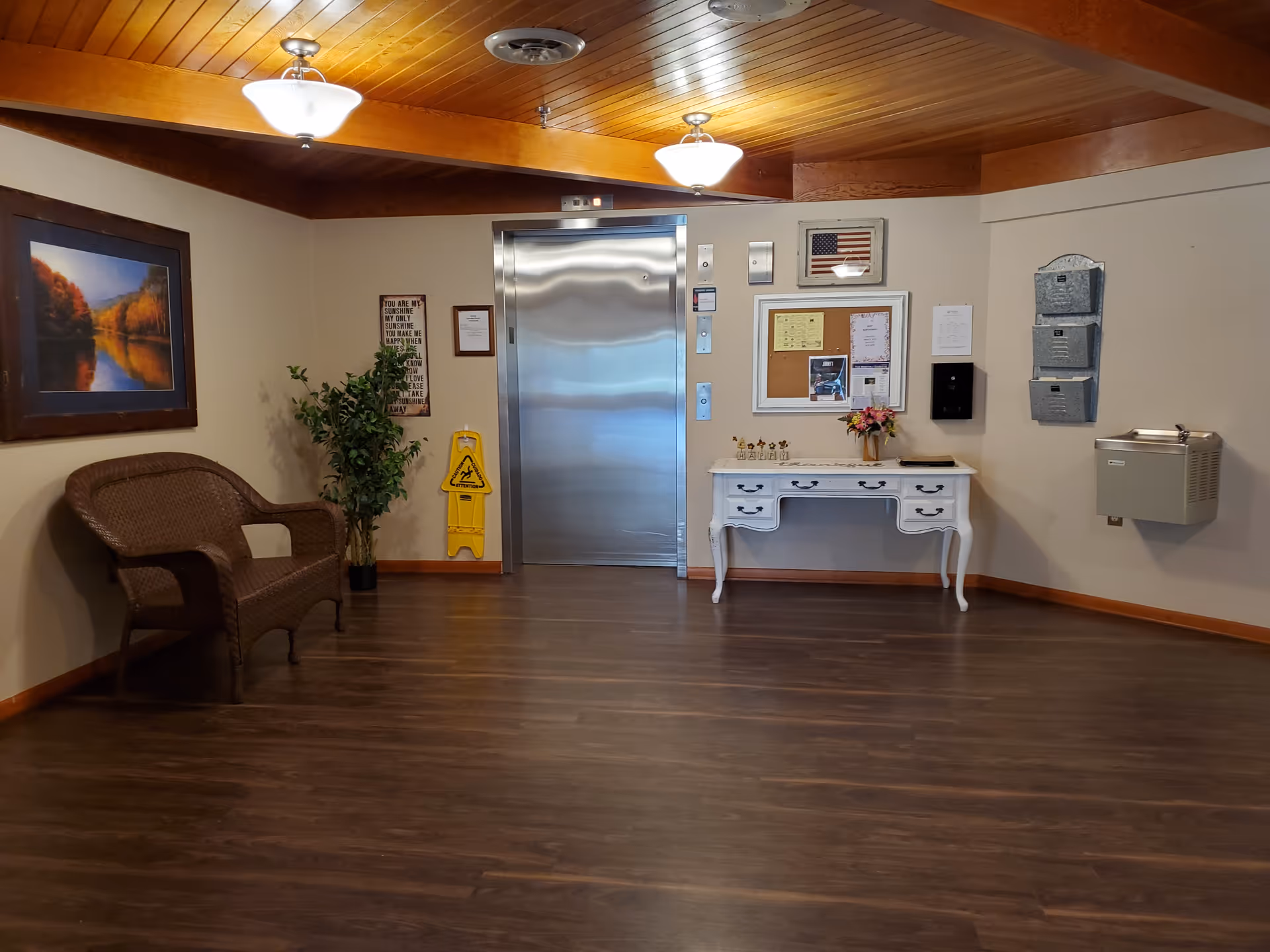 Small lobby area with an elevator, wicker bench, decorative table and bulletin board under wooden ceilings.