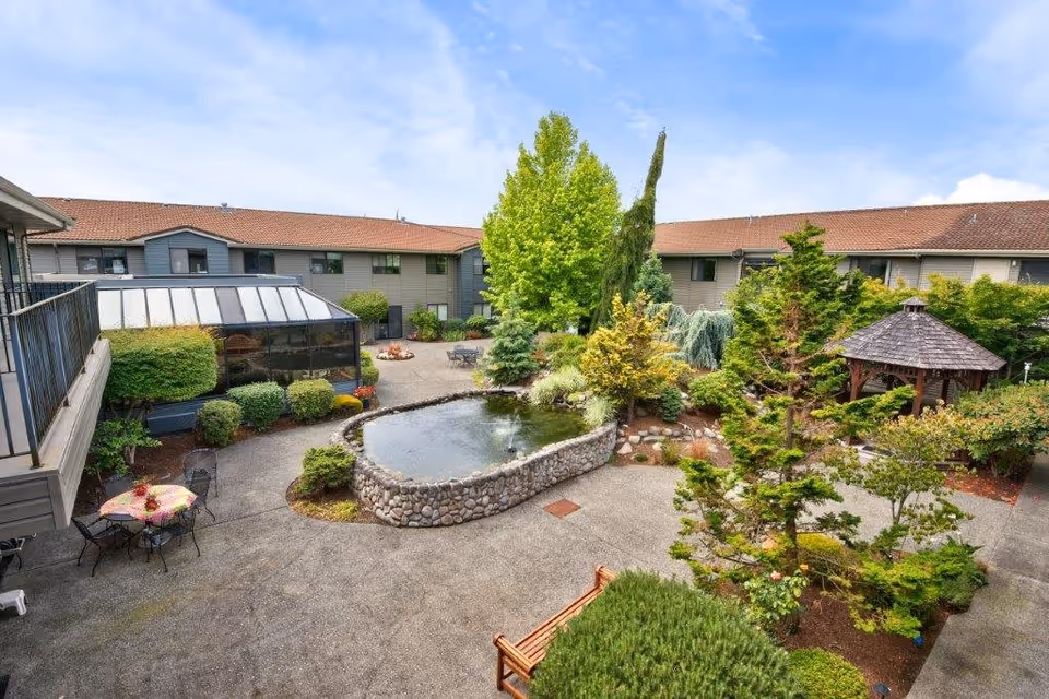 Outdoor courtyard area of Mountain Glen Retirement and Assisted Living featuring a stone-bordered pond with a small fountain, surrounded by trees, shrubs, and landscaped garden beds. There are several seating areas including metal tables and chairs with umbrellas, a wooden bench, and a gazebo with a shingled roof. The courtyard is enclosed by a two-story building with a red-tiled roof and gray siding under a partly cloudy blue sky.