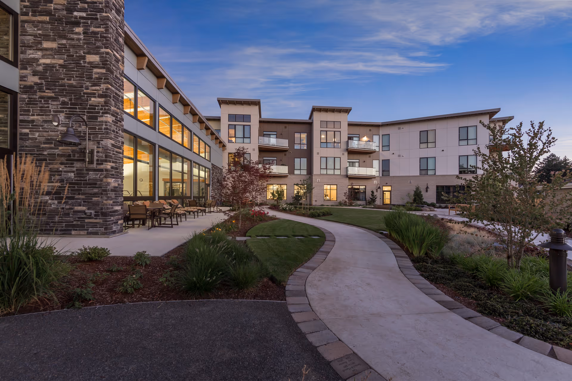 Exterior view of a senior living community building at dusk with a curved concrete pathway leading through landscaped gardens. The building has large windows, balconies, and outdoor seating areas with chairs and tables. The sky is clear with a soft blue hue.