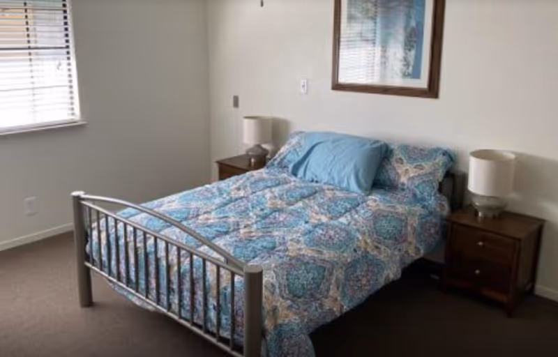 A bedroom with a metal frame bed covered with a blue patterned comforter and a blue pillow. There are two wooden nightstands on either side of the bed, each with a lamp. A window with blinds is on the left wall, and a framed picture hangs above the bed on the right wall.
