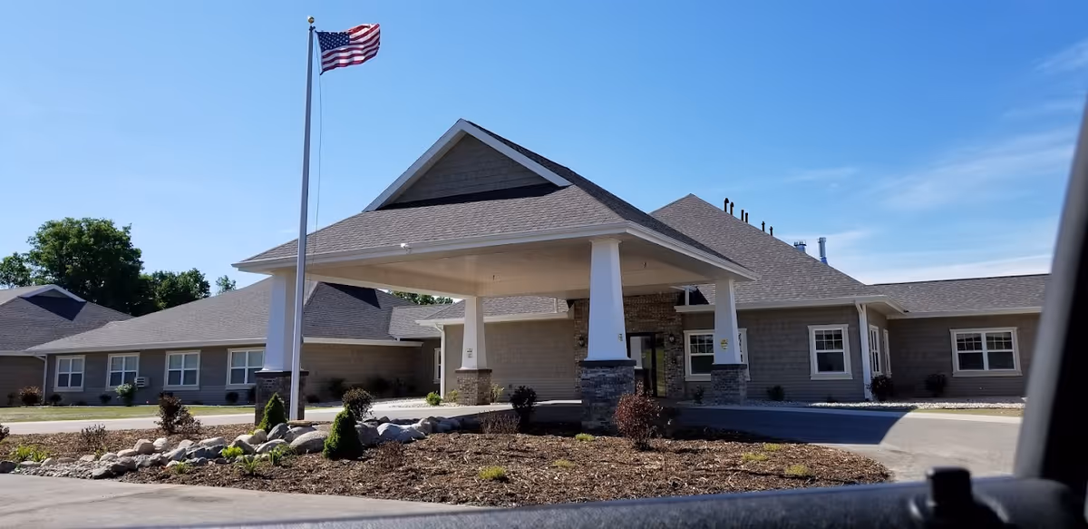 Exterior view of The Brook of Cedar Springs building with a covered entrance supported by white columns and an American flag on a flagpole in front. The building has gray siding, multiple windows, and a clear blue sky above.