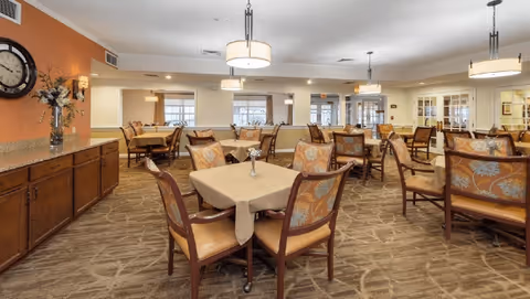 A spacious dining room with multiple tables covered with beige tablecloths and surrounded by wooden chairs with patterned cushions. The room has warm lighting from ceiling fixtures and wall sconces, a large clock on an orange accent wall, and windows letting in natural light.