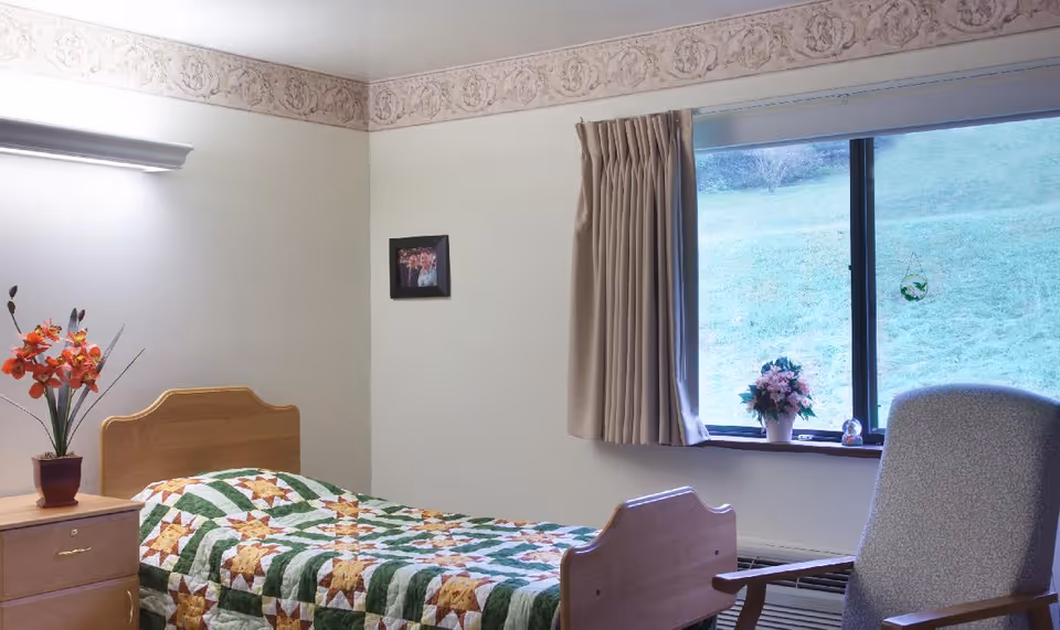 A simple bedroom in a senior living facility with a single wooden bed covered by a green, white, and brown star-patterned quilt. Next to the bed is a wooden nightstand with a vase of orange flowers. A large window with beige curtains shows a grassy outdoor area. A gray upholstered chair is positioned near the window. The walls are light-colored with a decorative border near the ceiling.