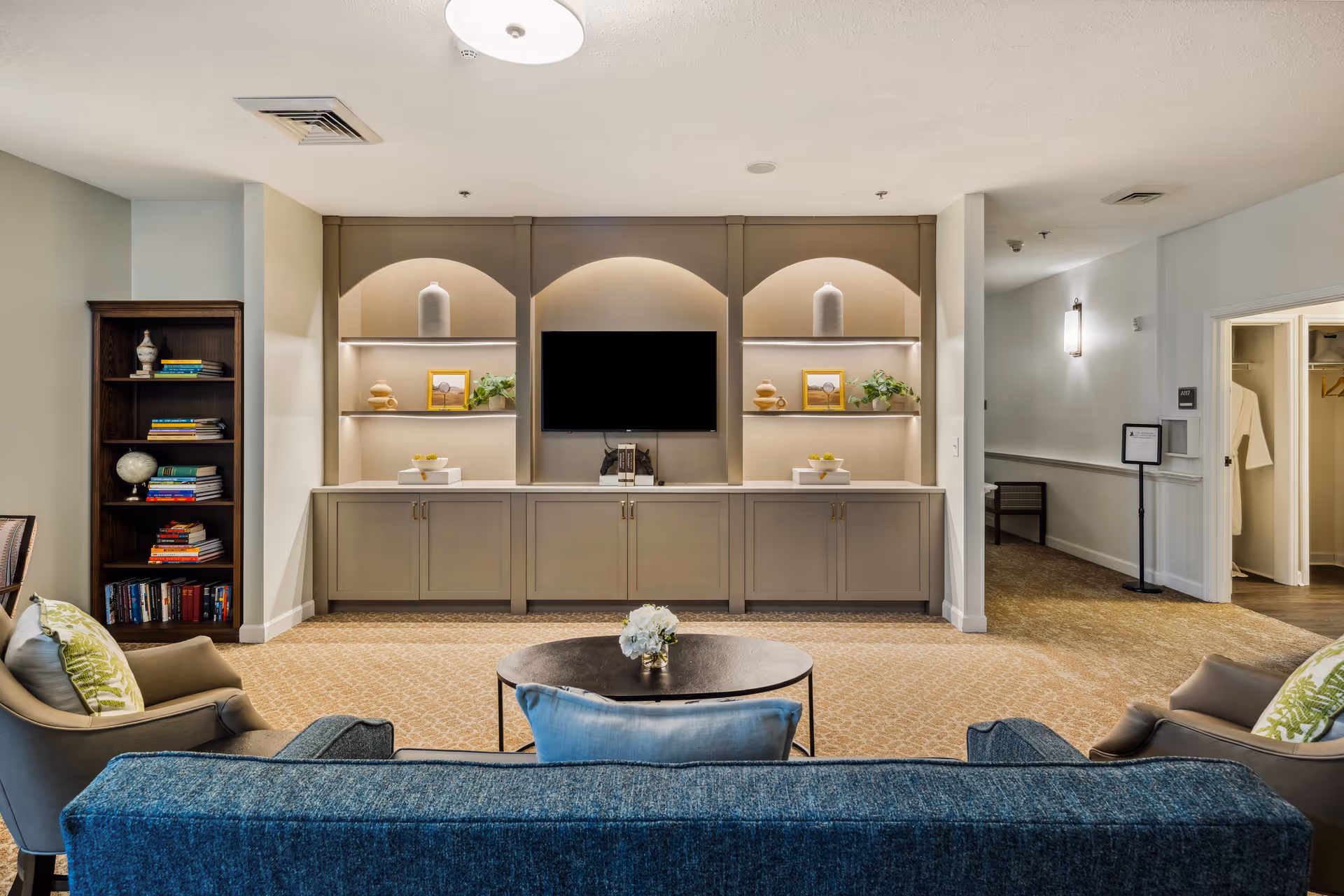 A cozy living room area with a blue sofa and two armchairs facing a built-in entertainment center with a flat-screen TV. The entertainment center has shelves with decorative items and framed pictures. There is a round coffee table with a small flower arrangement in front of the sofa. To the left, there is a tall bookshelf filled with books and decorative objects. The room has soft lighting and a beige carpeted floor.