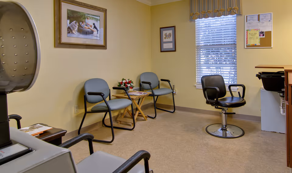 A small room with yellow walls and beige flooring, featuring three gray chairs with black armrests, a black salon chair, a wooden side table with magazines and a flower arrangement, a window with blinds and a valance, framed pictures on the walls, and a hair dryer chair in the foreground.