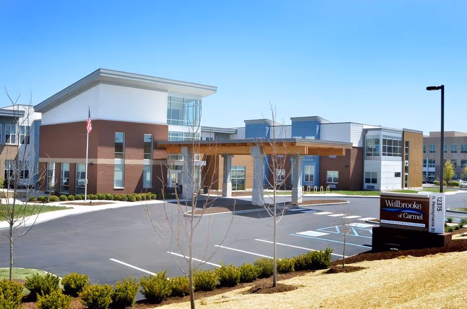 Exterior view of Wellbrooke of Carmel facility showing a modern two-story building with large windows, a covered entrance, an American flag, and a parking lot with marked spaces including handicapped spots. The sky is clear and blue, and there are small trees and shrubs in the landscaped area.