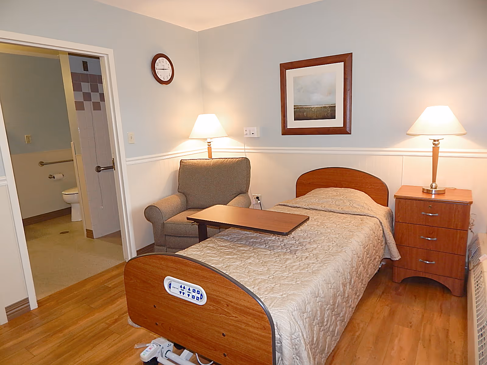 A senior living bedroom with a single adjustable bed covered with a beige quilt, a wooden nightstand with a lamp, an armchair with a small overbed table, a framed picture on the wall, and a clock. An open door reveals an accessible bathroom with grab bars and a toilet.