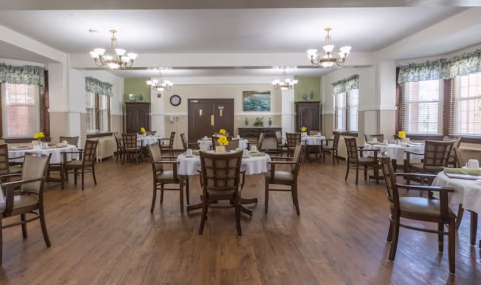 A spacious dining room with multiple round tables covered with white tablecloths, each set with cups, plates, and napkins. Wooden chairs surround the tables, and yellow flowers serve as centerpieces. The room has wooden flooring, large windows with floral valances, and chandeliers hanging from the ceiling. There are cabinets and a clock on the far wall.