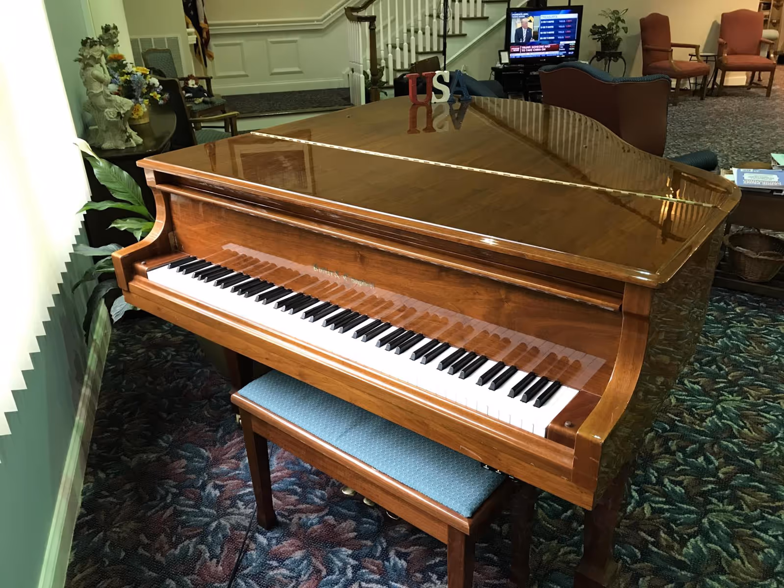 Polished wooden grand piano and bench in a carpeted lounge area with chairs and a TV in the background.