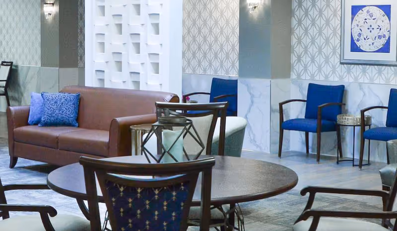 A supportive living facility common area featuring a brown leather sofa with blue pillows, several blue upholstered chairs with wooden arms, a round wooden table with decorative lanterns, and patterned wallpaper with framed artwork on the wall.
