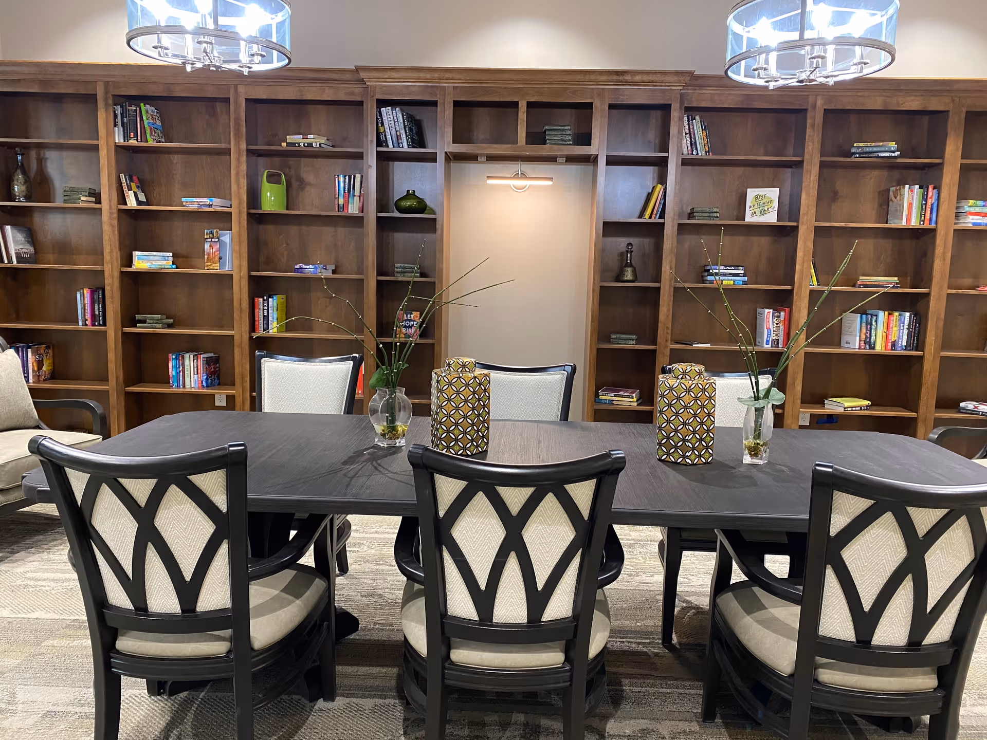 A communal library-style room with a large dark table and upholstered chairs in front of wooden bookshelves filled with books and decorative items.