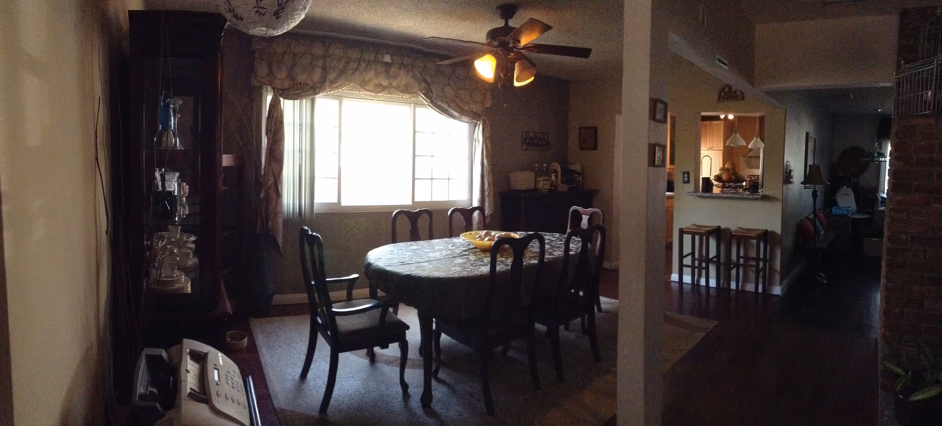 Dining room with an oval table covered by a tablecloth and six chairs under a ceiling fan, a china cabinet by a window, and an open view into the kitchen.