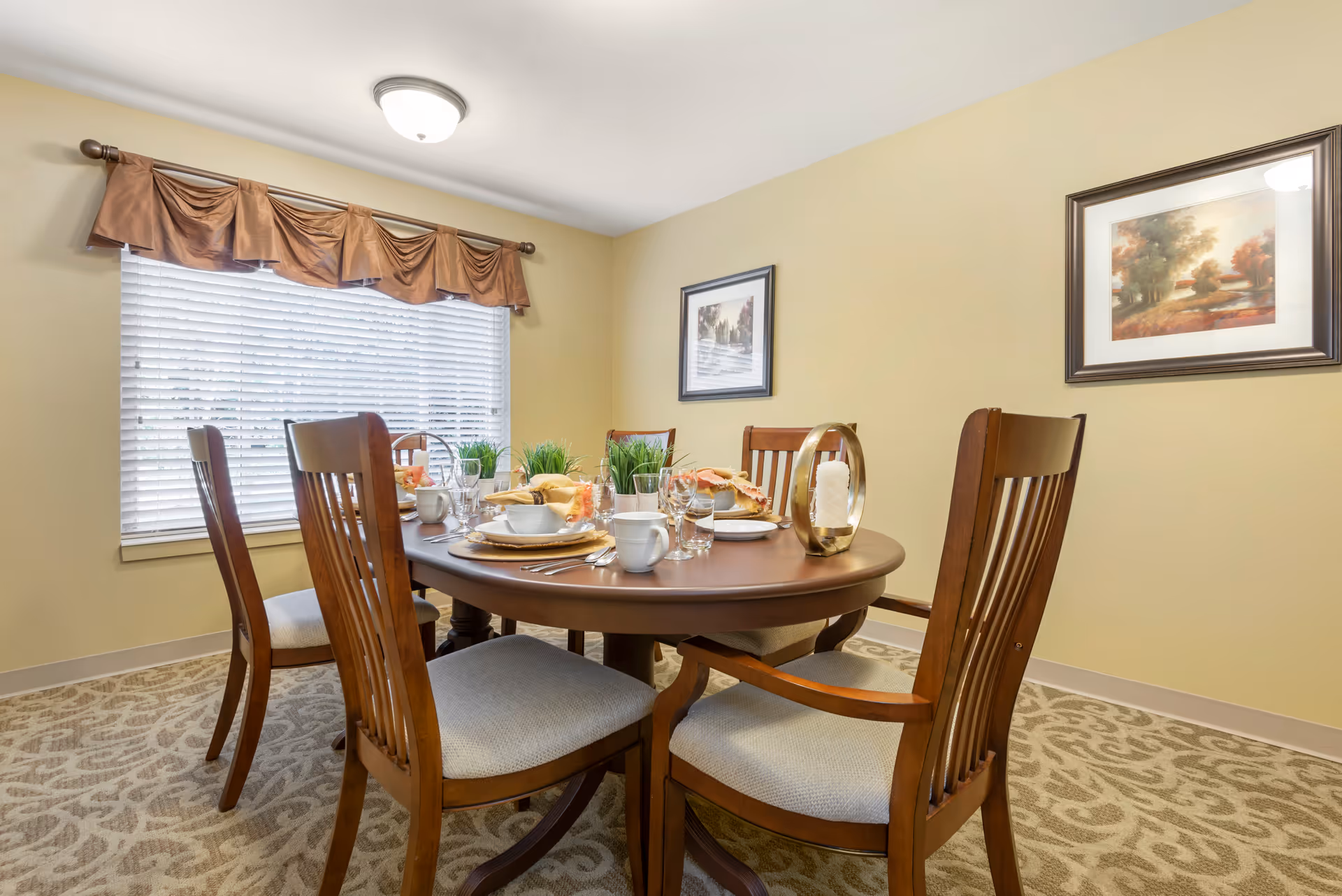 A cozy dining room with a round wooden table set for a meal, surrounded by wooden chairs, a window with blinds and a valance, and framed artwork on the walls.