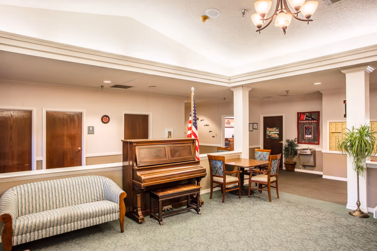 Interior common area of a senior living facility featuring a wooden upright piano with a bench, an American flag beside it, a striped upholstered sofa, a wooden table with four chairs, and several doors along the wall. The ceiling has recessed lighting and a chandelier, and there are plants and a water fountain in the background.