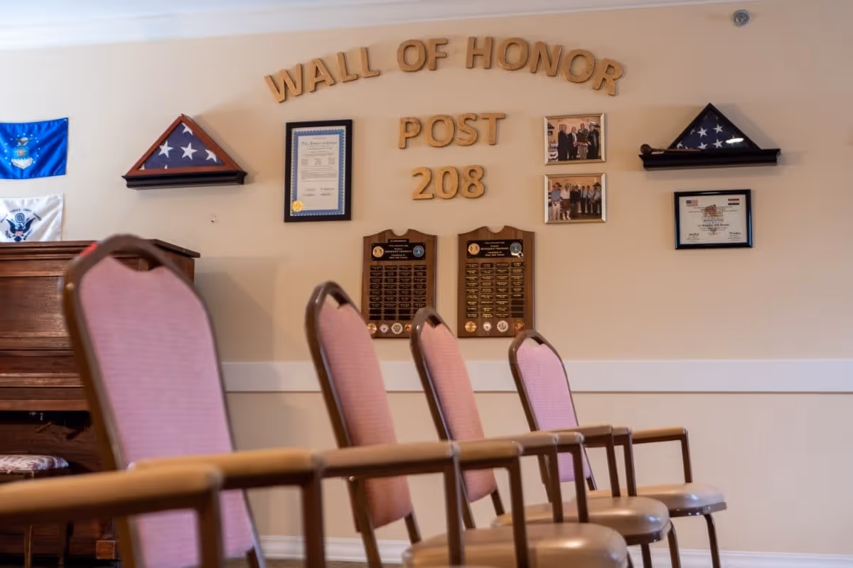 A room with a row of pink cushioned chairs facing a wall labeled 'Wall of Honor Post 208' with framed certificates, photographs, and folded American flags displayed on the wall.