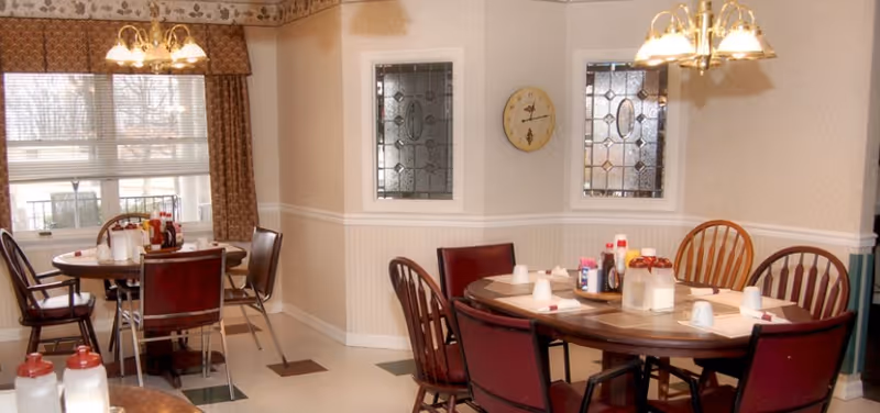 Interior view of a dining room with two round wooden tables surrounded by chairs. Each table has place settings with cups, napkins, and condiments. The room features patterned wallpaper, two decorative stained glass windows, a wall clock, and hanging light fixtures. A large window with curtains allows natural light into the space.