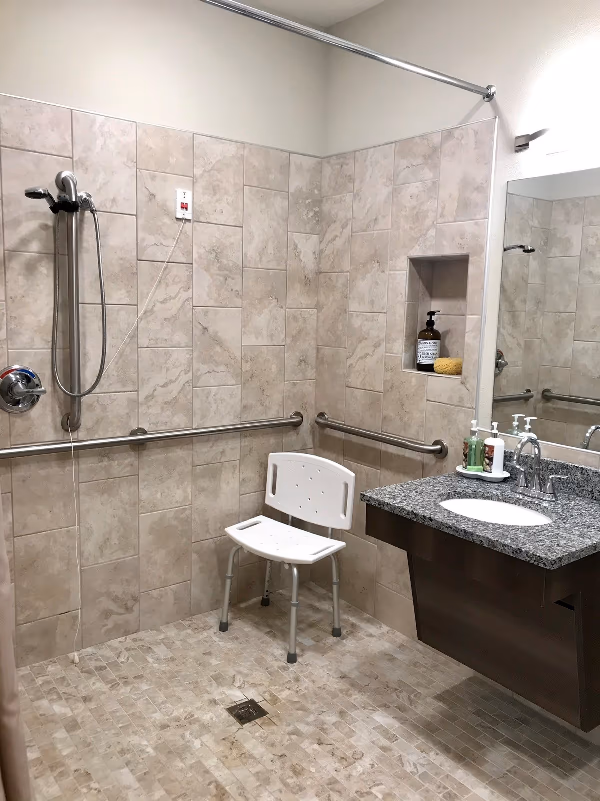 Accessible bathroom with beige tiled walls and floor, a white shower chair, grab bars along the walls, a handheld showerhead, and a granite countertop with a sink and soap dispensers. There is a recessed shelf in the wall holding a bottle and a sponge.