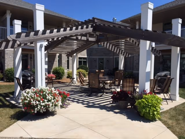 Outdoor courtyard with a wooden pergola shading a patio dining table and chairs, flanked by potted flowers in front of a two-story building.
