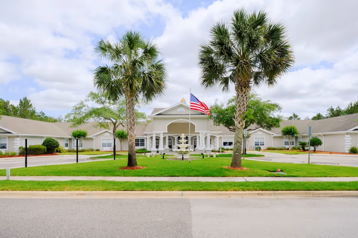 Front exterior view of Grand Villa of Palm Coast building with a central entrance featuring white columns, an American flag on a flagpole, a tiered fountain, two palm trees, and well-maintained green lawn and landscaping under a partly cloudy sky.
