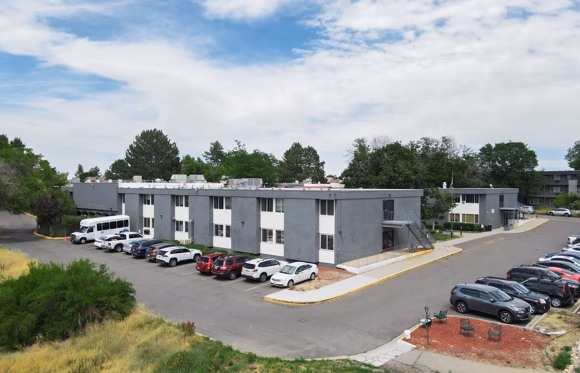 Exterior view of Cherrelyn Health Care Center showing a two-story gray and white building with multiple windows, surrounded by parked cars and greenery under a partly cloudy sky.
