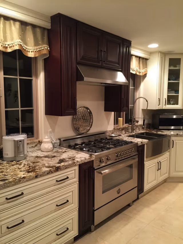 Modern kitchen with granite countertops, stainless steel gas stove and oven, a large farmhouse sink, dark wood and cream cabinetry, two windows with beige valances, and various kitchen appliances on the counter.