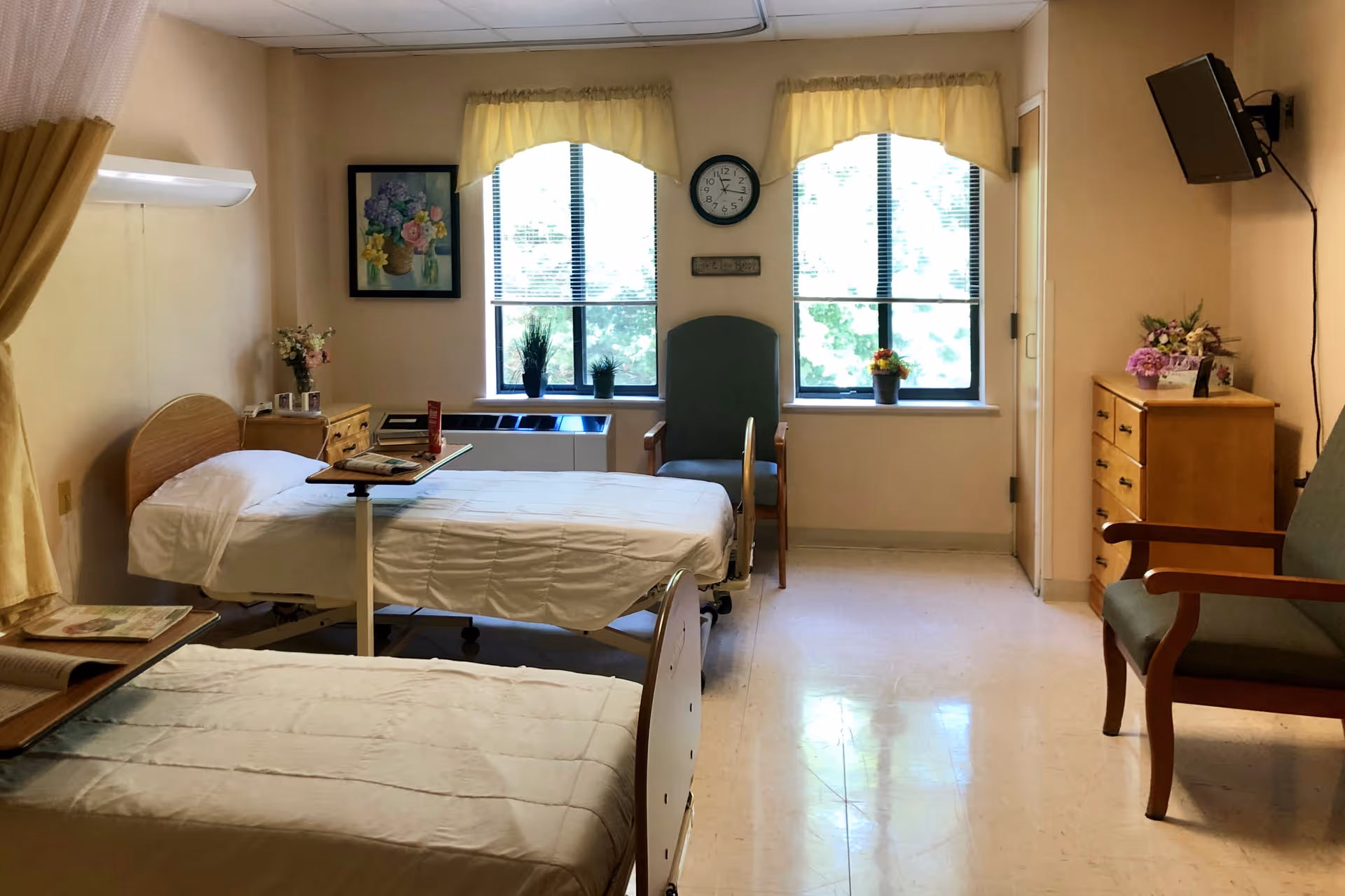 A bright and clean room with two hospital-style beds, each with white bedding and an adjustable overbed table. The room has two large windows with yellow valances, a clock on the wall between the windows, and a framed floral painting. There are two green armchairs, a wooden dresser with flowers on top, and a wall-mounted television in the corner.