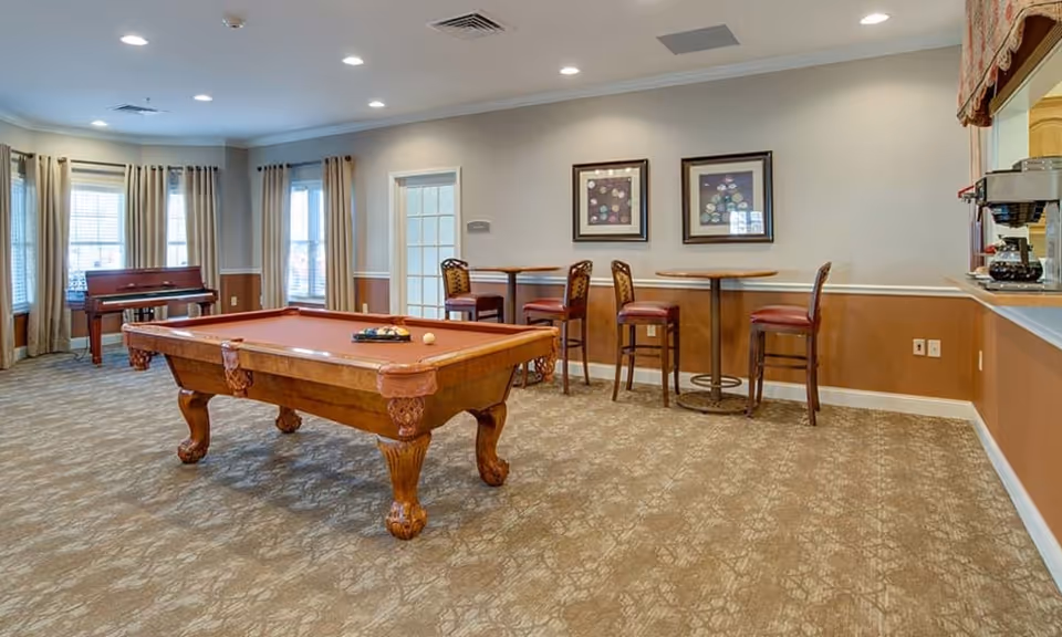 A spacious room with a pool table in the center, two round tables with chairs against the wall, a piano near the windows with beige curtains, and a coffee station on the right side. The room has carpeted flooring and neutral-colored walls with framed artwork.