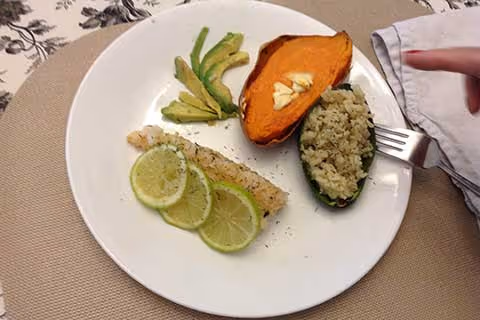 A white plate with a meal consisting of a piece of fish topped with three lime slices, sliced avocado, half a baked sweet potato with garlic cloves, and a stuffed jalapeño pepper with rice. A fork is placed on the right side of the plate on a beige placemat.