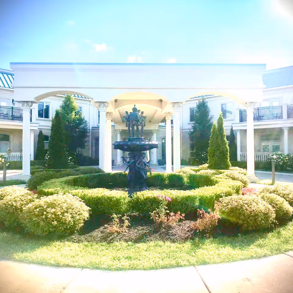 Front entrance of a senior living building with a central decorative fountain, trimmed hedges, and a columned portico.