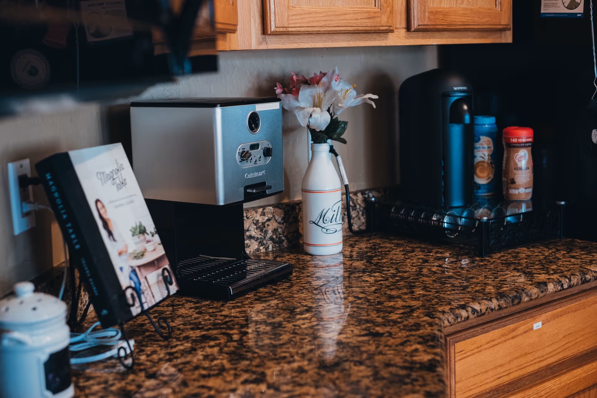 A kitchen countertop with a coffee maker, a milk bottle vase with flowers, coffee pods in a tray, and a cookbook on a stand. Wooden cabinets are visible above and below the granite countertop.