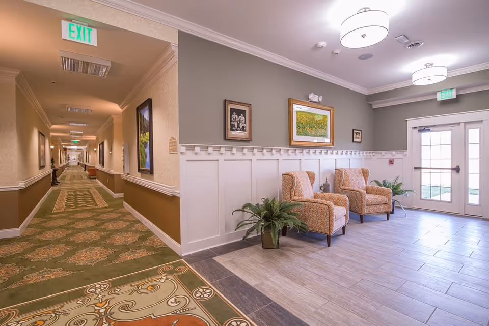 A well-lit hallway and sitting area in a senior living facility. The hallway has patterned green and brown carpeting with framed pictures on the walls. The sitting area features two patterned armchairs, potted plants, and framed artwork on the walls. Double glass doors with exit signs are visible, letting in natural light.