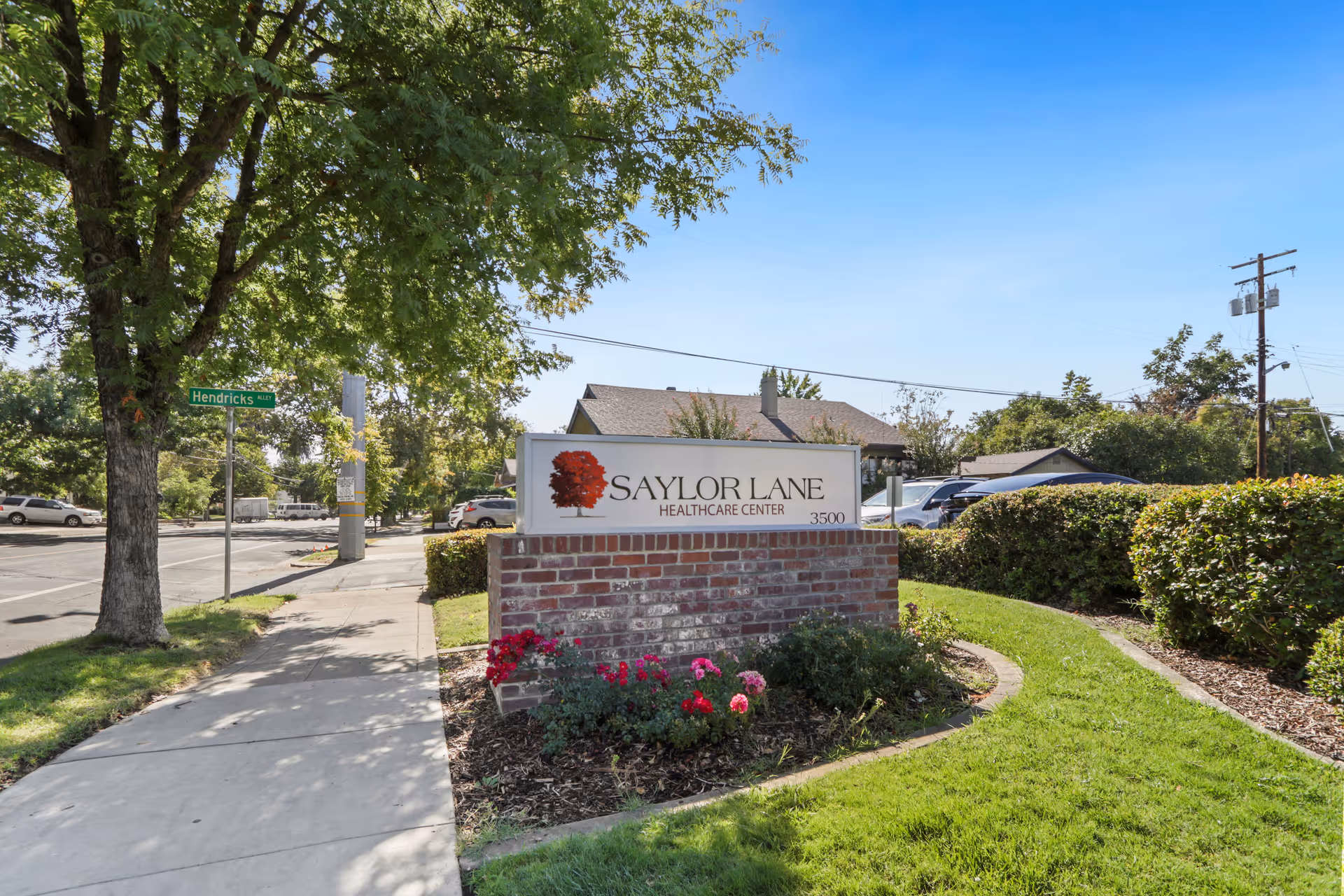 Outdoor view of the entrance sign for Saylor Lane Healthcare Center, mounted on a brick base with flowers and greenery around it. The scene includes a sidewalk, a tree, street signs, parked cars, and a clear blue sky.