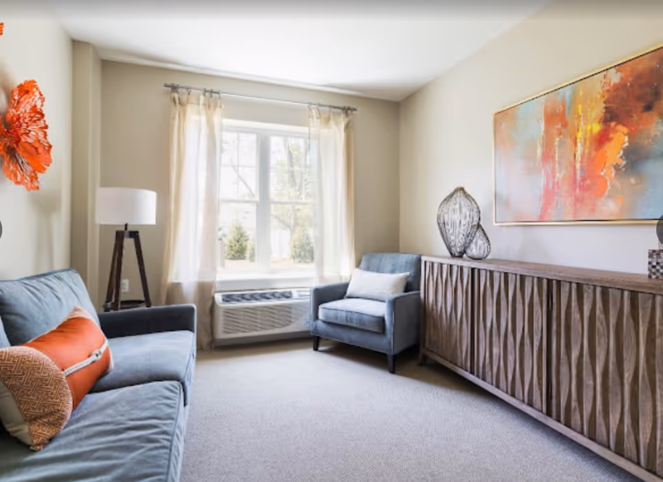 A bright living room with a large window covered by sheer curtains. The room features a blue sofa with orange and patterned pillows, a matching blue armchair with a white pillow, a wooden sideboard with a textured front, decorative glass vases, a colorful abstract painting on the wall, and a floor lamp with a white shade.