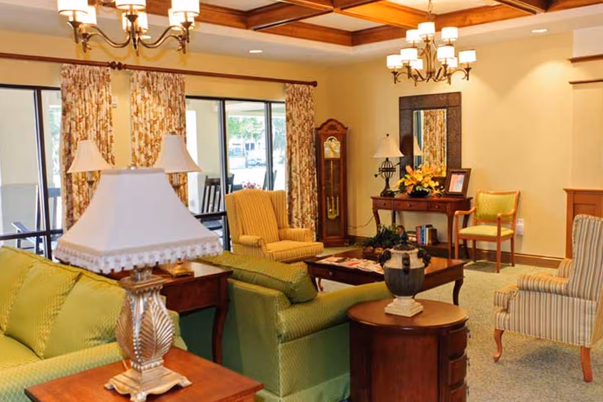 A cozy living room with green and striped armchairs, a green sofa, wooden side tables with lamps, a wooden coffee table with decorative items, a grandfather clock, a console table with a mirror and flowers, and floral curtains covering large windows.